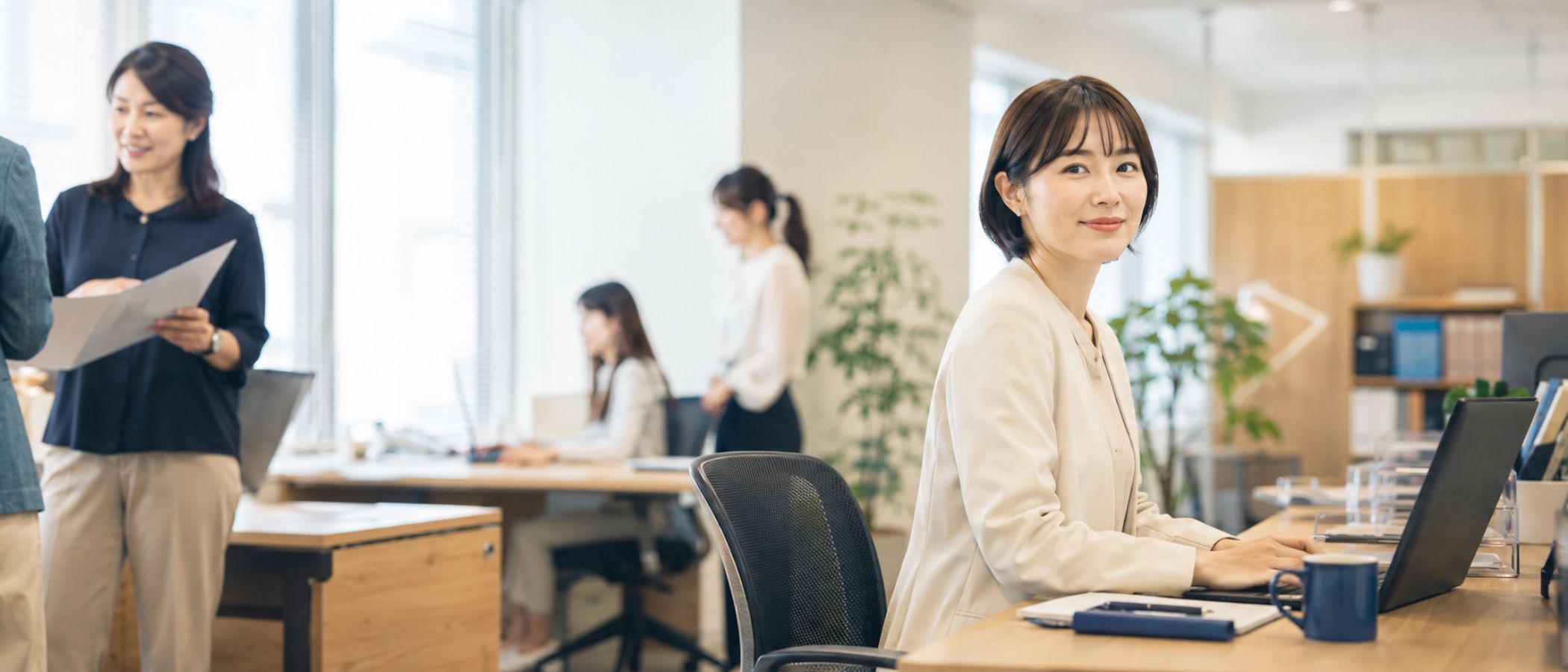 窓辺のやわらかな自然光に包まれたオフィスで、ノートとPCとグリーンが整えられた事務派遣比較サイトのメインビジュアル2